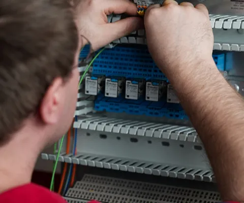 A technician in a red uniform is working on an electrical control panel, adjusting components and wiring inside a cabinet.