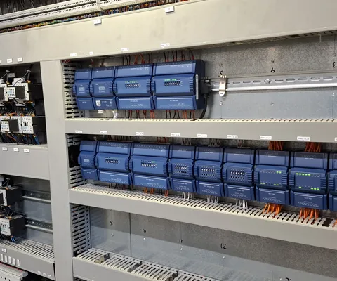 Een bedieningspaneel met blauwe elektronische modules en oranje bedrading, georganiseerd op metalen rekken, dat een complexe elektrische installatie laat zien.