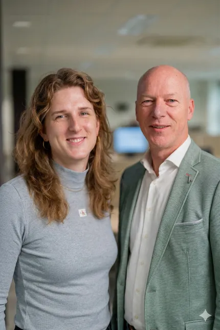 Two people smiling in an office setting. One wears a gray turtleneck, the other a light green blazer and white shirt. Blurred background.