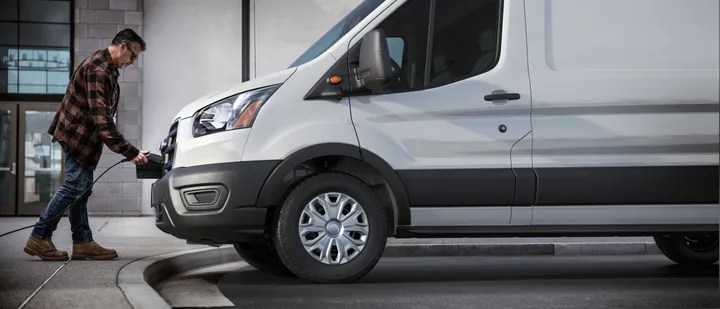 Man in plaid shirt plugs charger into white van parked near a building.