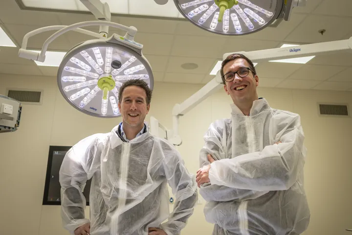 Projectmanager Danny van der Put en lead engineer Arie Kraaijeveld lachen terwijl ze gekleed in beschermende kleding in een behandelruimte van het LUMC LIGT-centrum staan