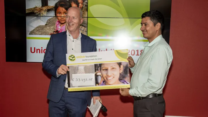 Two men smiling, holding a large donation check for €33,492.45 from the Unica Foundation, in front of a screen displaying foundation visuals.