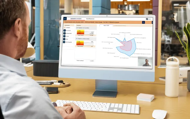 Man sitting at a desk using a computer displaying analytical charts and a video conference call, surrounded by office items.