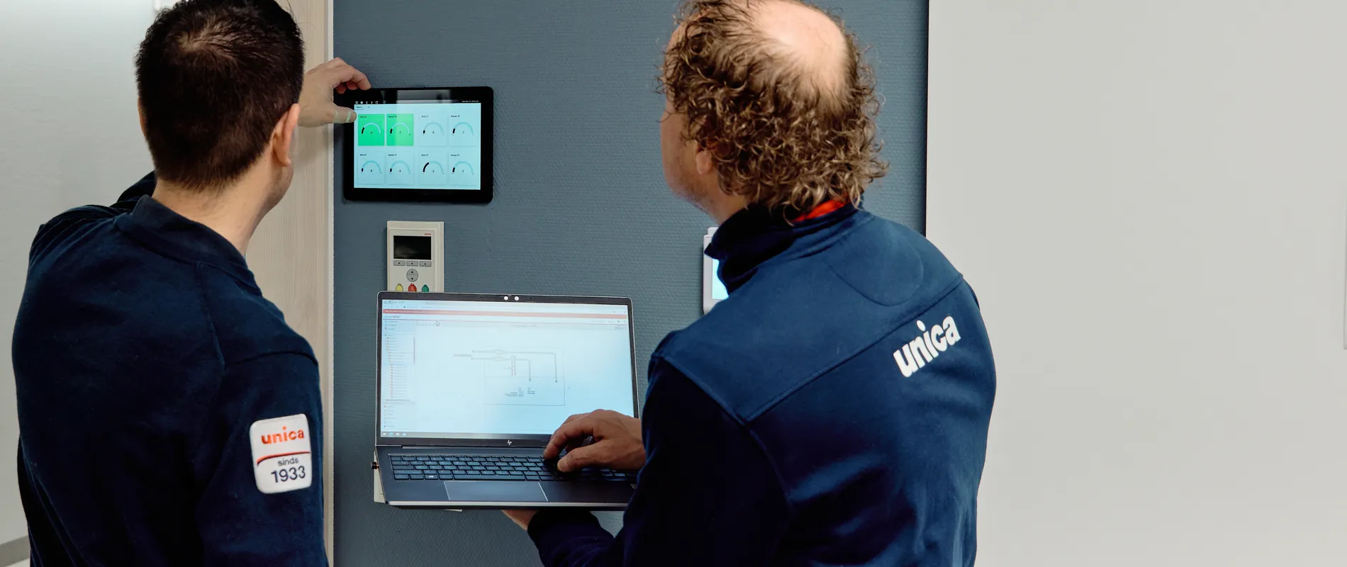 Twee medewerkers van Unica checken de status van de regeltechnische installatie via wandpaneel en laptop
