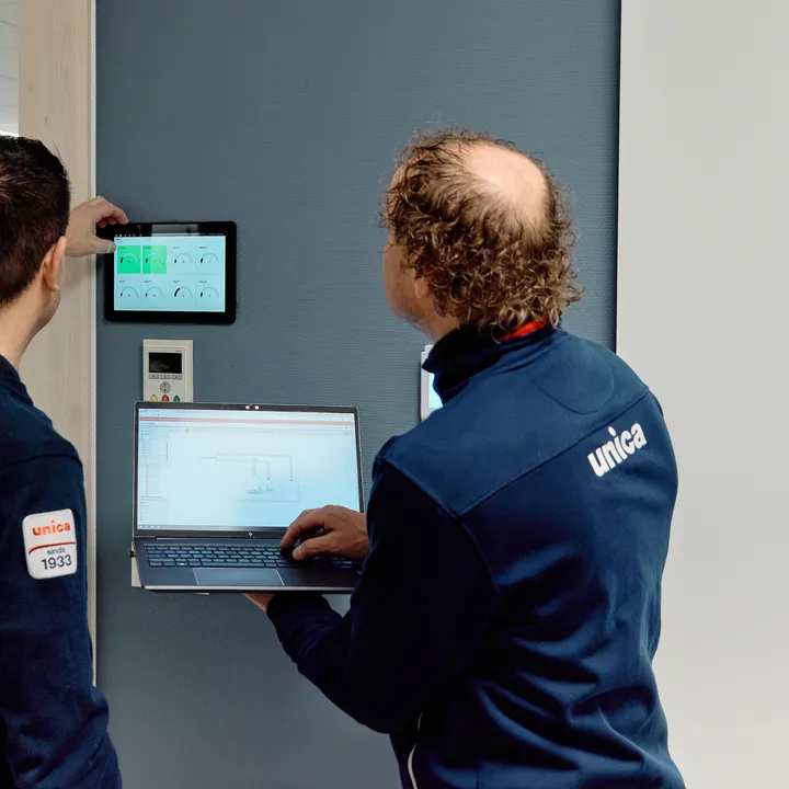 Twee medewerkers van Unica checken de status van de regeltechnische installatie via wandpaneel en laptop