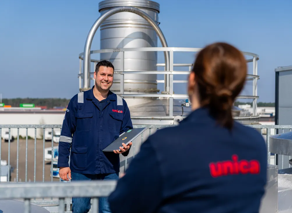 Twee Unica-technici in gesprek op het dak van een industriële installatie met koeltoren op de achtergrond