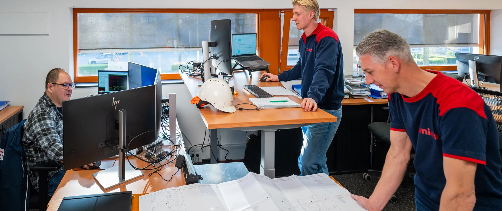 Team van engineers bekijkt tekeningen en werkt aan werkplekken in een technisch kantoor.