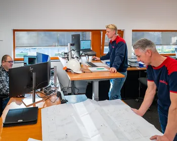 Team van engineers bekijkt tekeningen en werkt aan werkplekken in een technisch kantoor.