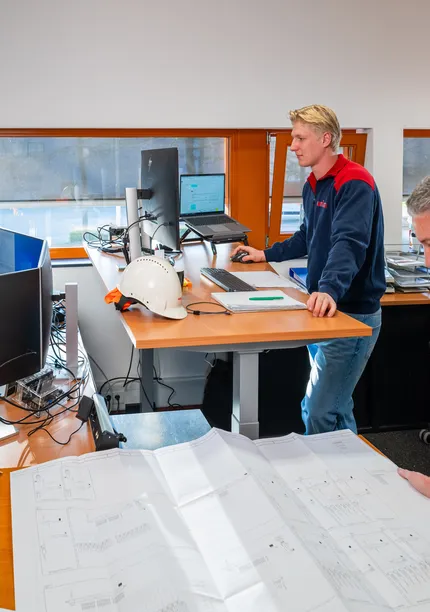 Team van engineers bekijkt tekeningen en werkt aan werkplekken in een technisch kantoor.