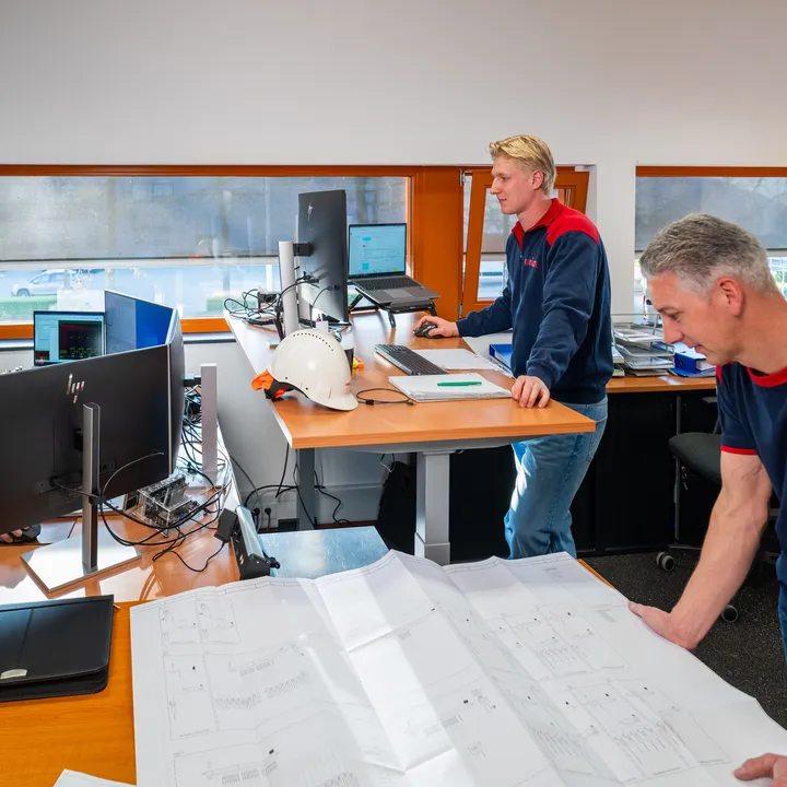 Team van engineers bekijkt tekeningen en werkt aan werkplekken in een technisch kantoor.