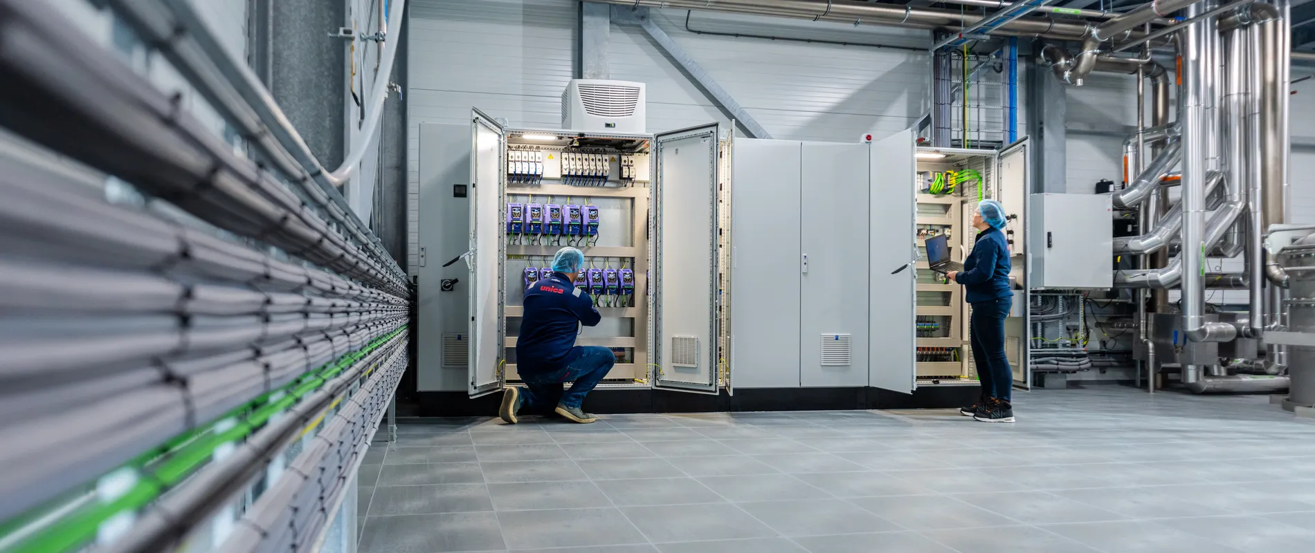 Twee medewerkers inspecteren open schakelkasten; de een werkt met een laptop, de ander voert bedrading na.
