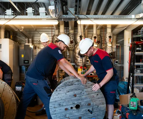 Workers in hard hats and uniforms maneuver a large wooden cable spool in an industrial setting with tools and equipment around.