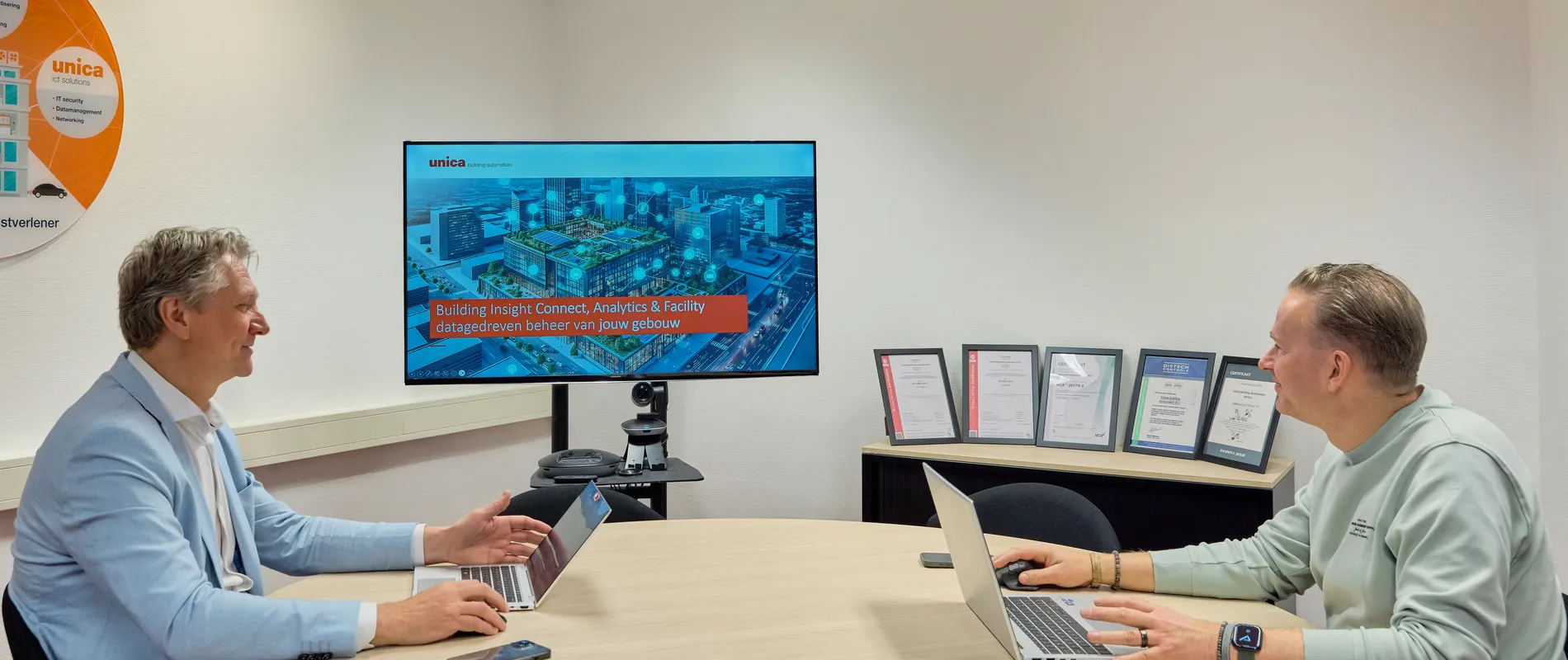 Twee lachende mensen tegenover elkaar aan tafel, beide met laptops opengeklapt en een beeldscherm met presentatie op de achtergrond.