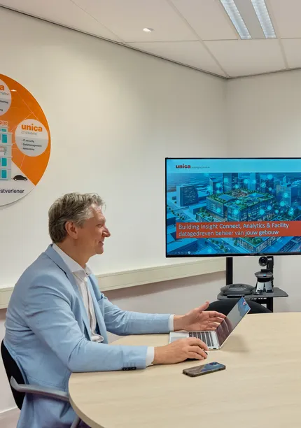 Twee lachende mensen tegenover elkaar aan tafel, beide met laptops opengeklapt en een beeldscherm met presentatie op de achtergrond.