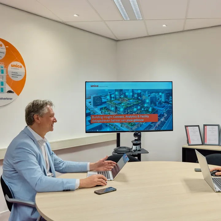 Twee lachende mensen tegenover elkaar aan tafel, beide met laptops opengeklapt en een beeldscherm met presentatie op de achtergrond.
