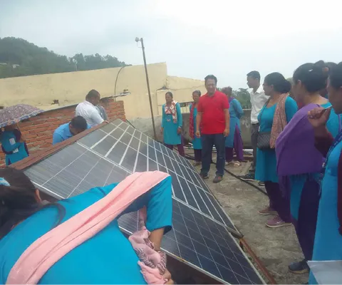 People gathered around solar panels, with women in blue uniforms and a man in a red shirt. A hilly background is visible.