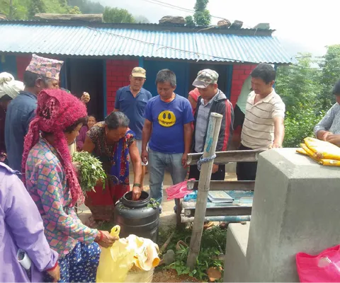 A group of people gather around a woman pouring liquid into a container outdoors, with a blue-roofed building in the background.