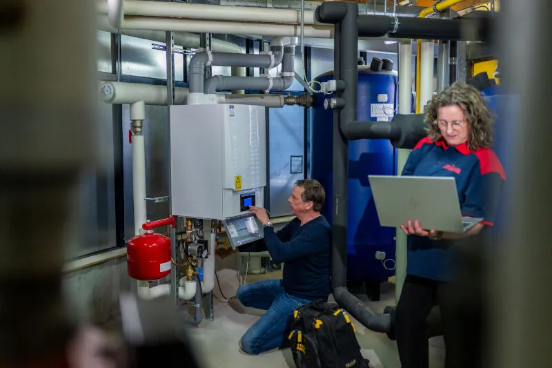Technicians in a boiler room; one adjusts a control panel, another uses a laptop. Industrial pipes and equipment surround them.
