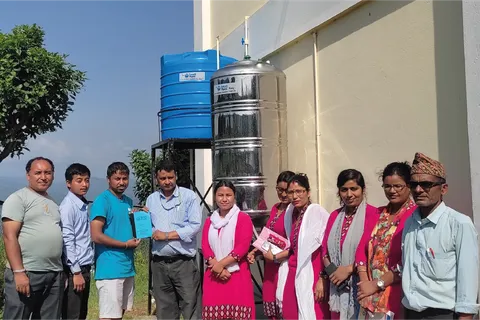 A group of people standing next to large blue and silver water tanks outside a building, holding documents, with trees and sky in the background.