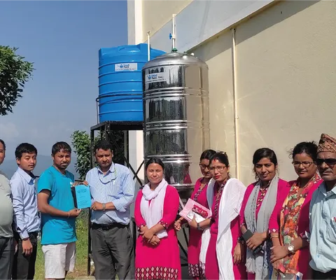 A group of people standing next to large blue and silver water tanks outside a building, holding documents, with trees and sky in the background.