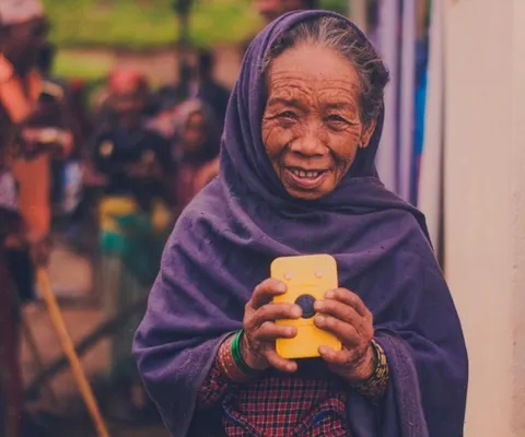 Elderly woman in a purple shawl, smiling and holding a yellow device. Background shows a blurred outdoor setting with people.