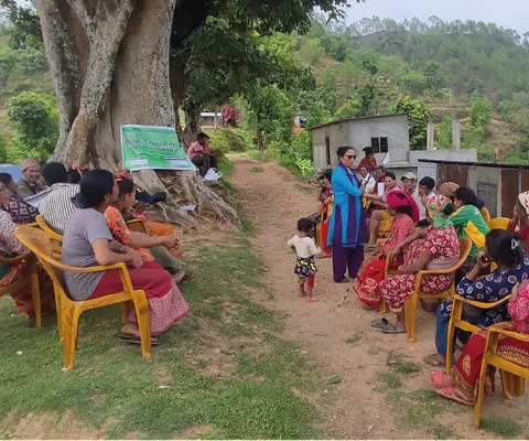 A group of people sit in a circle of chairs under large trees in a rural area, with a woman and child standing in the center.
