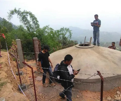 People standing near a large, dome-shaped concrete water tank on a hillside, surrounded by trees and a barbed wire fence.