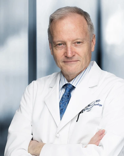 Dr. John Marshall, a doctor in a white coat and blue tie stands with arms crossed in front of a blurred background.