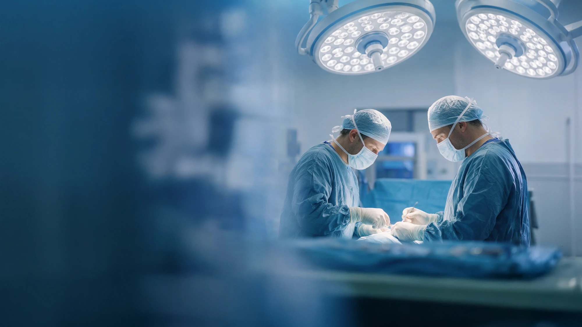 Two surgeons in blue scrubs and masks perform surgery under bright operating lights in a sterile hospital environment.
