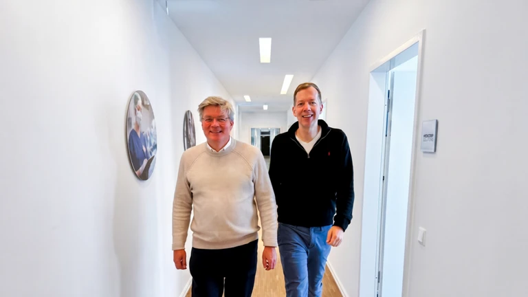 Two men smiling while walking down a bright hallway with wooden floors and white walls adorned with circular photos.
