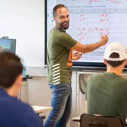 Ein Lehrer der Wilhelm-Bläsig-Schule unterrichtet drei Rehabilitanden im Klassenraum und trägt Formeln in eine Tabelle ein