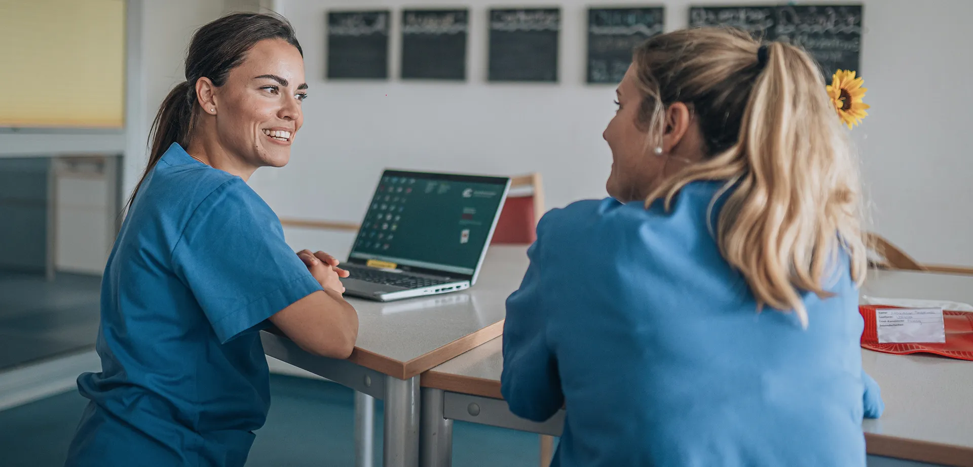 Zwei Mitarbeitende aus der Pflege in Arbeitskleidung im Austausch an einem LAptop