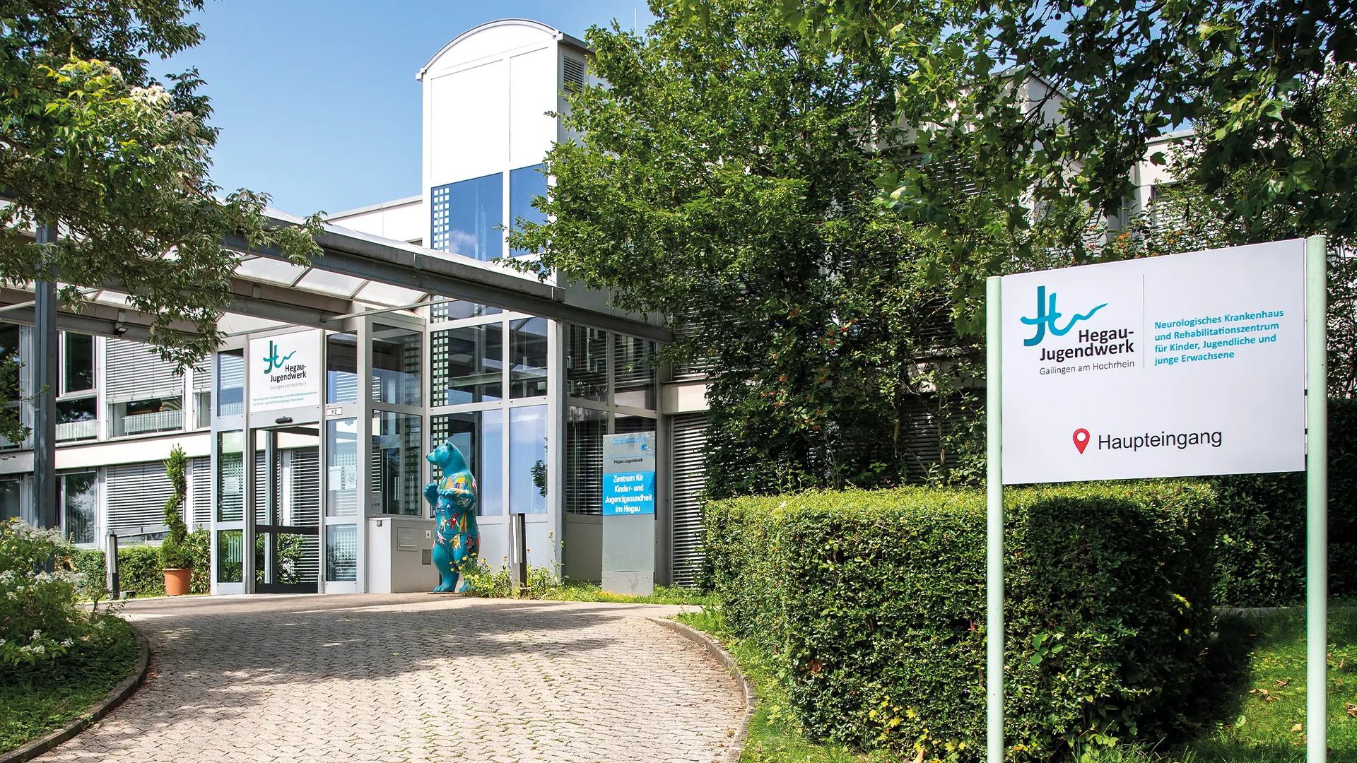 Haupteingang des Hegau-Jugendwerks in Gailingen mit dem Standort-Schild ‚Hegau-Jugendwerk‘ und einer bunten Bärenfigur rechts vom Eingang.