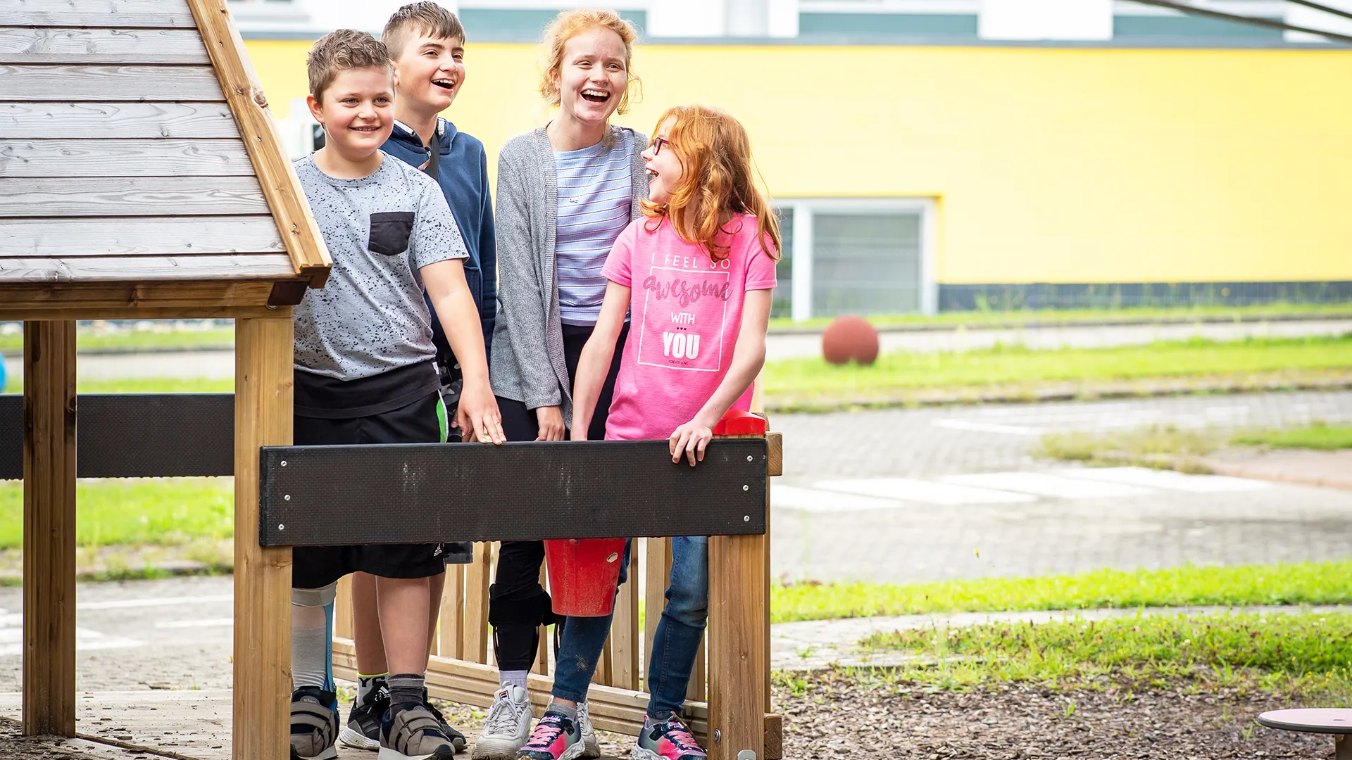 Zwei junge Rehabilitandinnen und zwei junge Rehabilitanden stehen als Gruppe neben einem Holzhaus auf dem Spielplatz des Hegau-Jugendwerks und lachen gemeinsam.