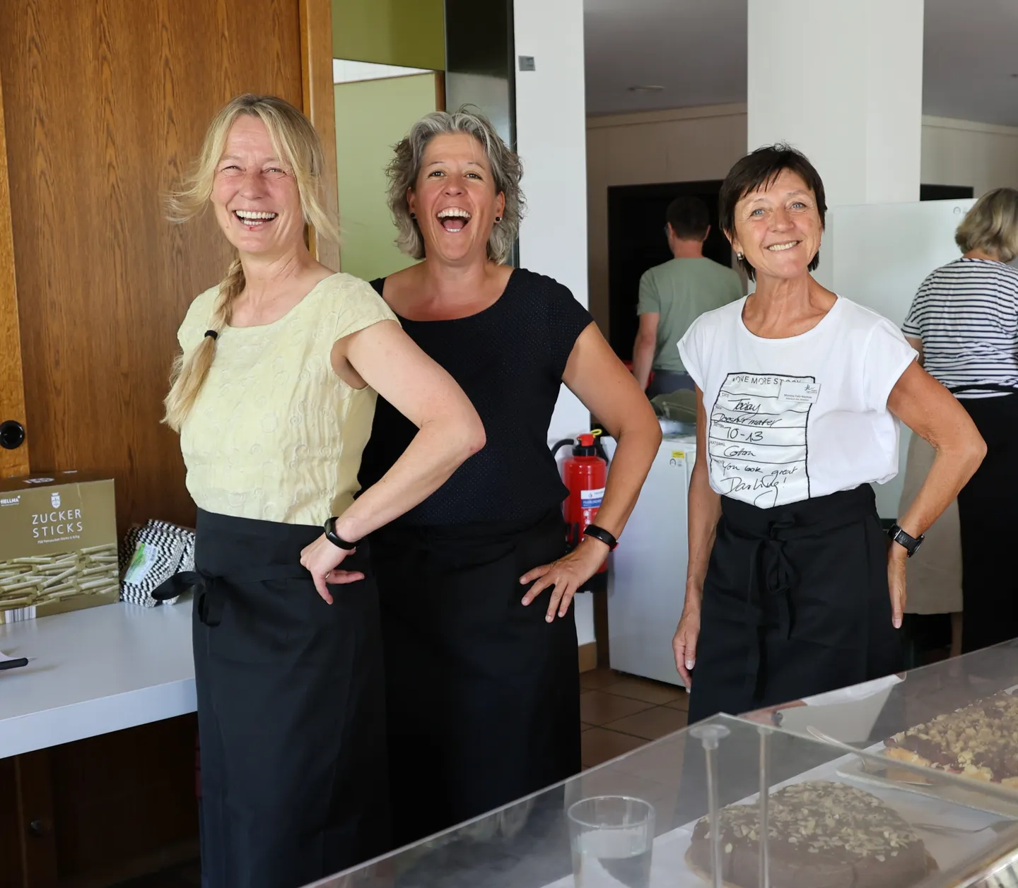 Three women wearing aprons smile and pose in a kitchen setting, with desserts on a counter and a fire extinguisher in the background.