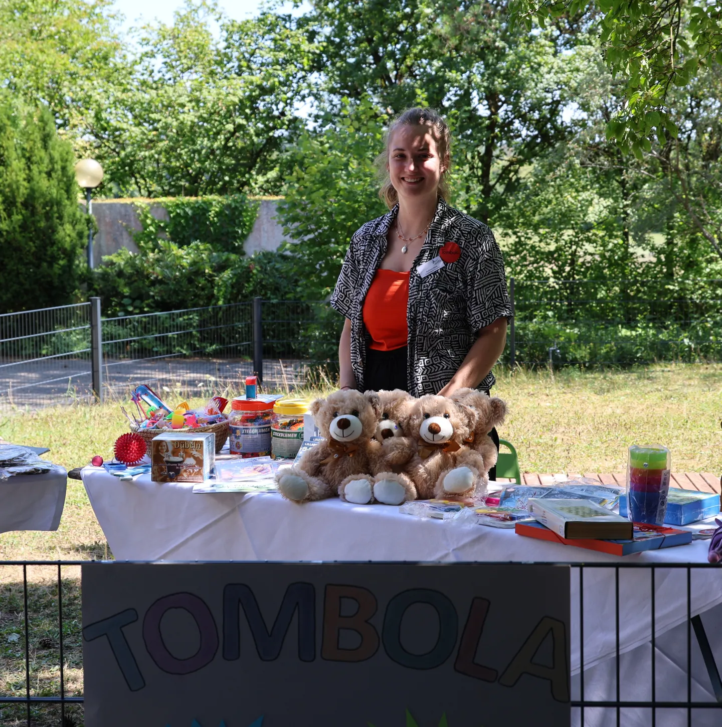 A woman stands behind a table with teddy bears and various items on display at an outdoor tombola event.