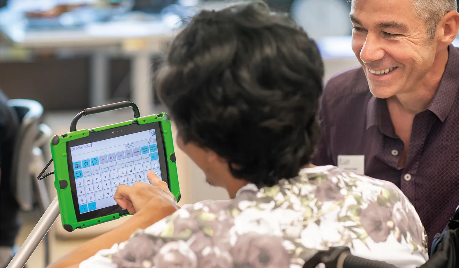 Pädagoge der Klinikschule sitzt mit einem jungen Rehabilitanden vor einem iPad als Hilfsmittel der Unterstützten Kommunikation. Der Rehabilitand tippt einen Satz auf dem Bildschirm