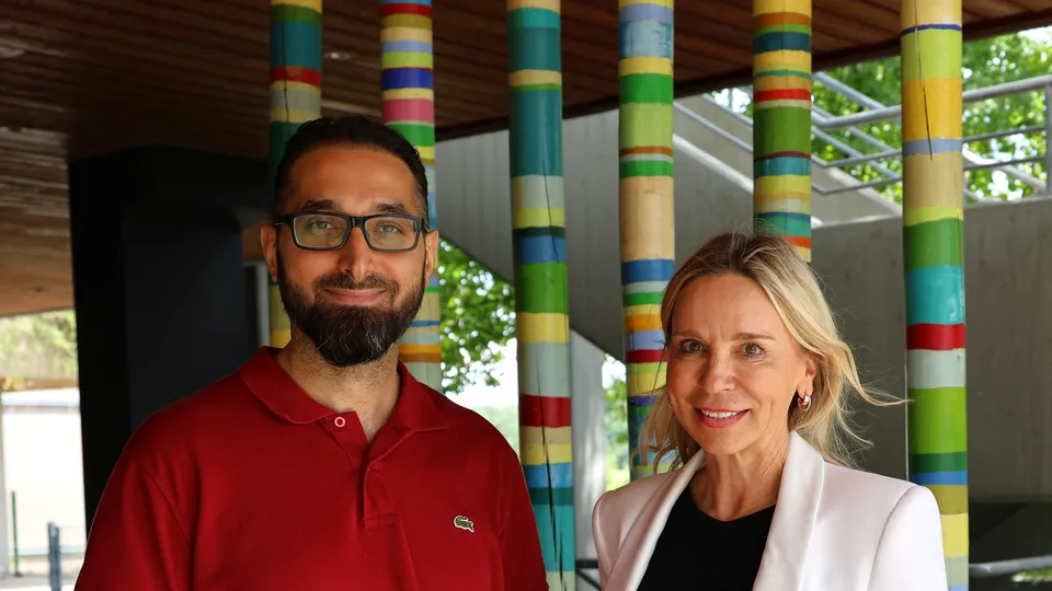 A man in a red polo shirt and a woman in a white suit stand smiling in front of colorful striped pillars.
