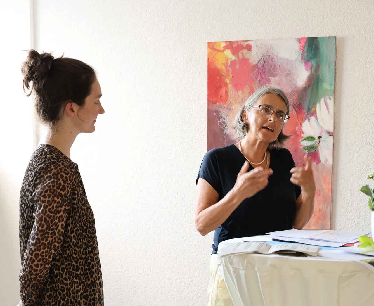Two women talking in front of a colorful abstract painting; one gesturing, the other listening attentively.