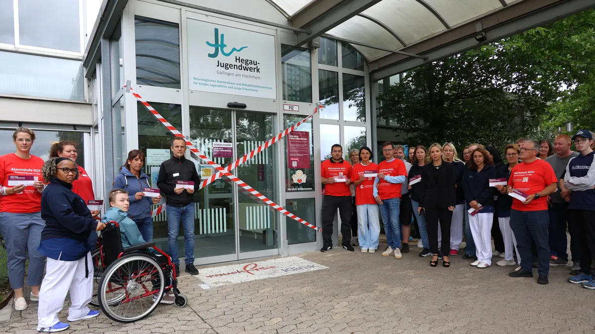 A group of people stand with a wheelchair user in front of a building with crossed tape and a "Hegau-Jugendwerk" sign.
