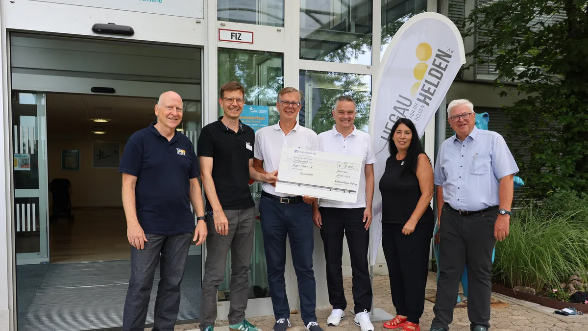 Six people stand in front of a building holding a large check, smiling at the camera.