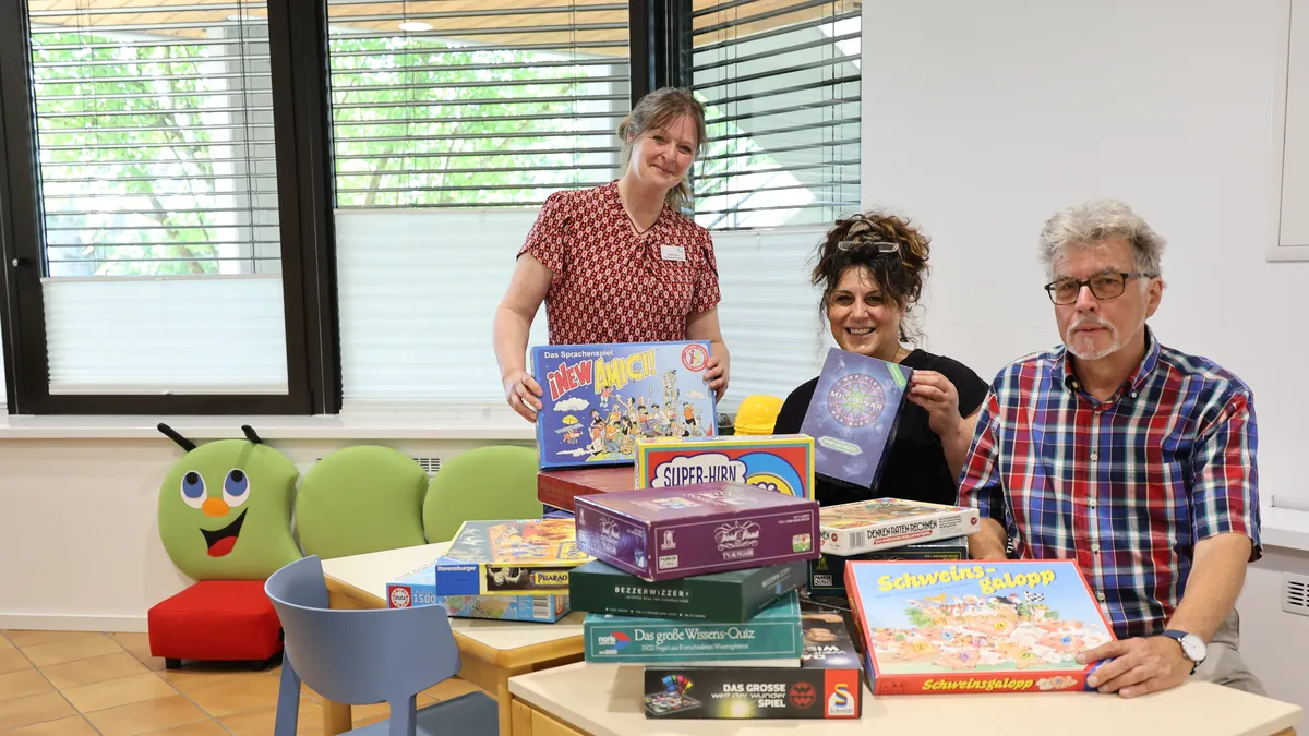 Three people sitting at a table with various board games, smiling and displaying game boxes in a bright room with large windows.