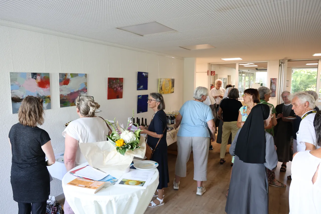 People gather in a hallway at an art exhibit, observing paintings on the wall and socializing near a table with flowers and refreshments.