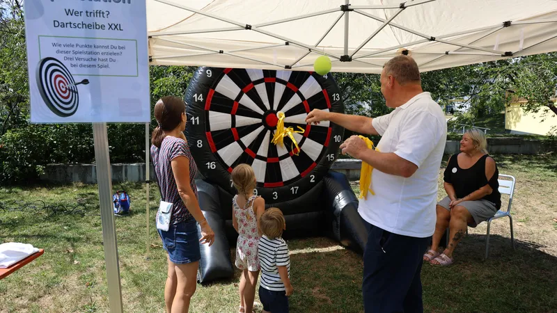 People playing with a giant inflatable dartboard under a tent. A man is throwing darts while children and a woman watch.