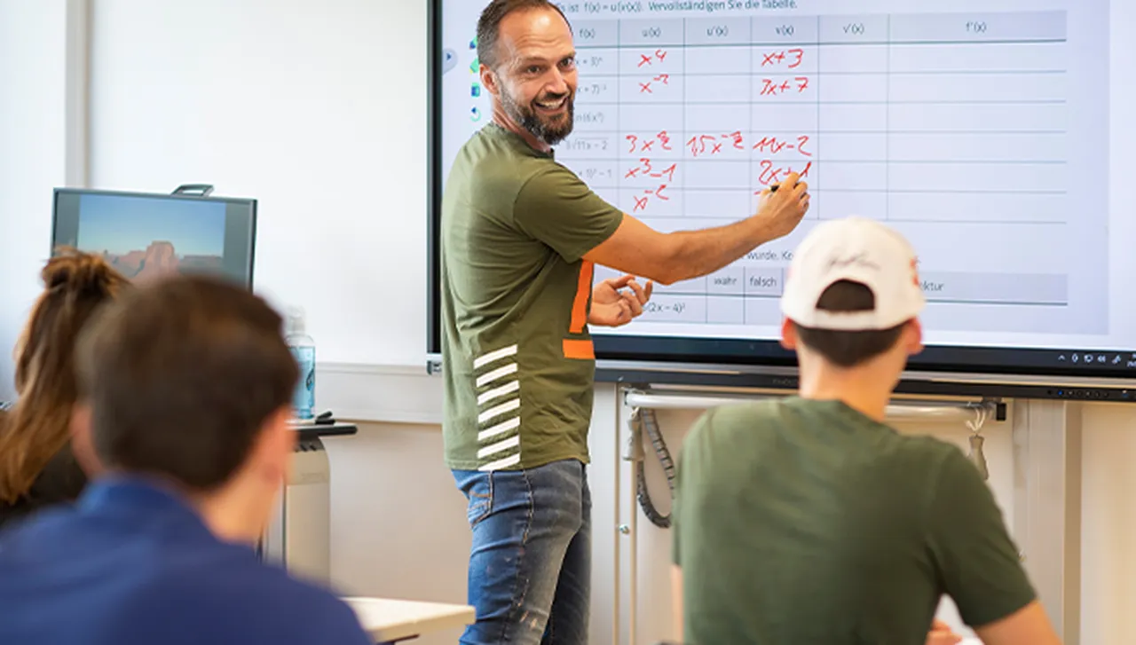 Regelschulgruppe in der Klinikschule im Hegau-Jugendwerk: Eine Schülergruppe arbeitet gemeinsam mit einem Lehrer im Unterricht, während dieser am Whiteboard schreibt und sich den Schülerinnen und Schülern zuwendet.