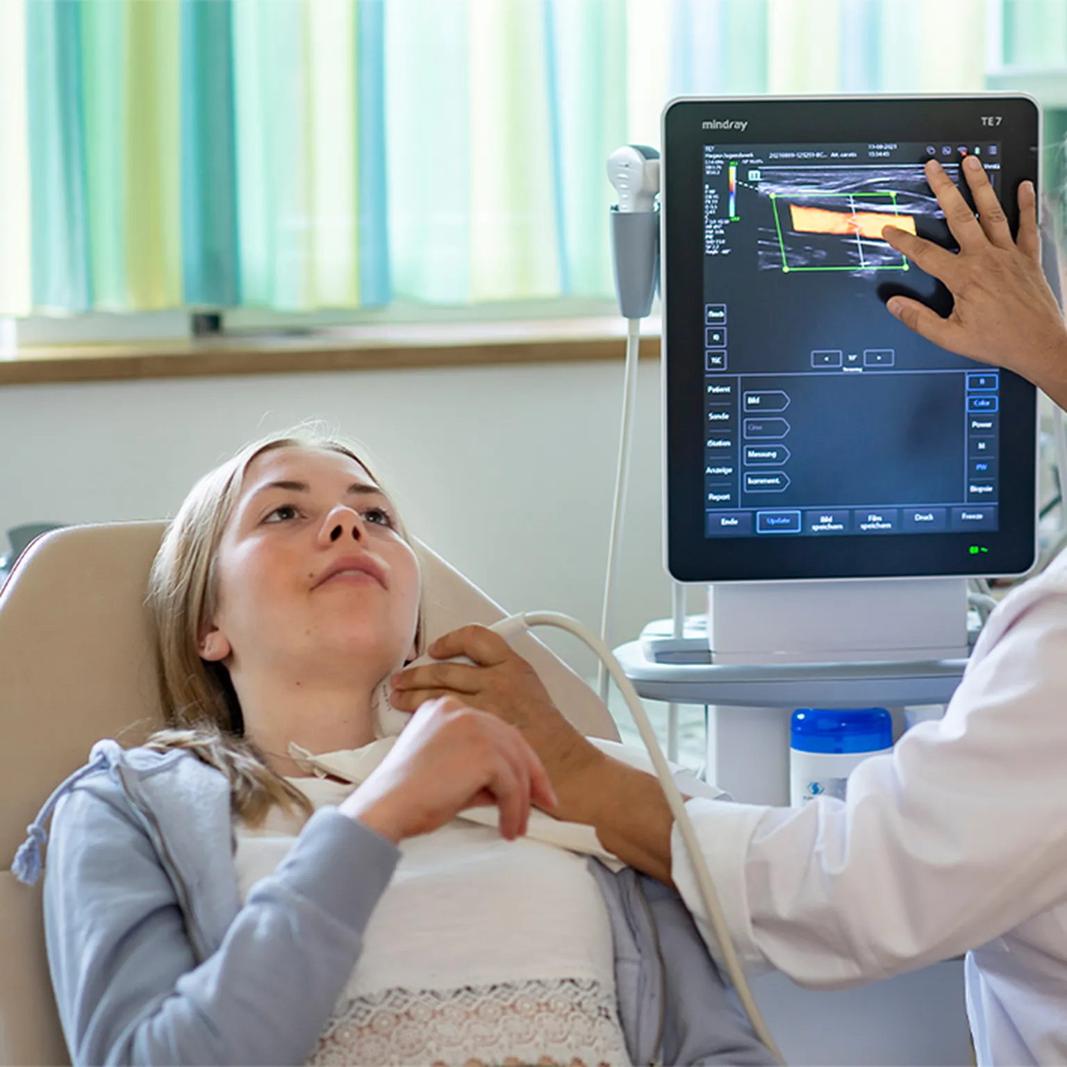 Junge Rehabilitandin liegt während einer Ultraschalluntersuchung des Halses auf einer Liege. Eine Ärztin führt den Schallkopf am Hals der Rehabilitandin an. Auf einem Monitor ist das Ultraschallbild zu sehen.
