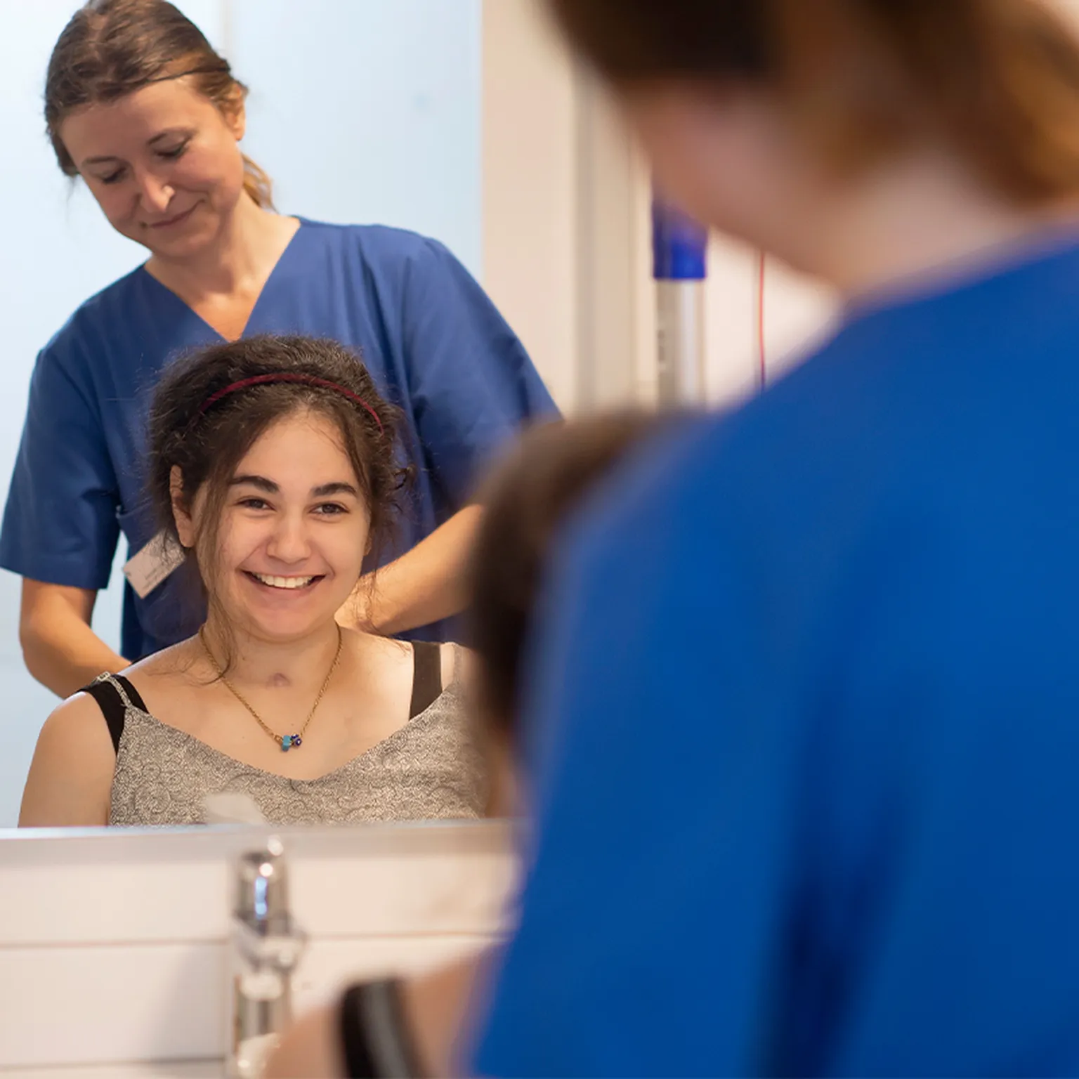Eine Pflegekraft kämmt einer jungen Rehabilitandin die Haare. Das Gesicht der Rehabilitandin ist im Spiegel zu sehen. Die Szene vermittelt pflegerische Unterstützung und persönliche Begleitung im Hegau-Jugendwerk.