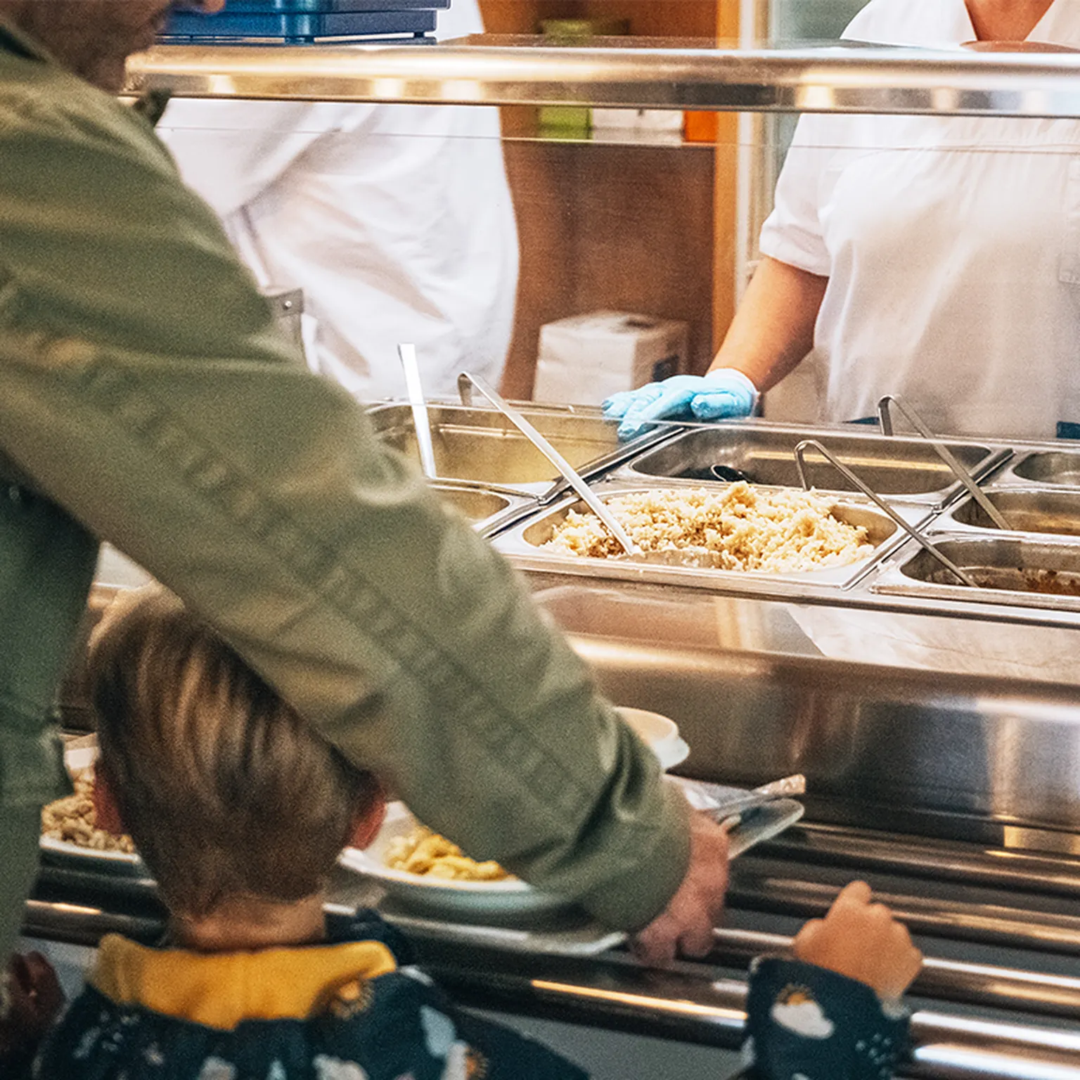 Ein Mann steht mit seinem Sohn vor der Essensausgabe in einer Cafeteria des Hegau-jugendwerks. Beide sind von hinten zu sehen. Hinter der Theke befindet sich eine Person, die Essen ausgibt.