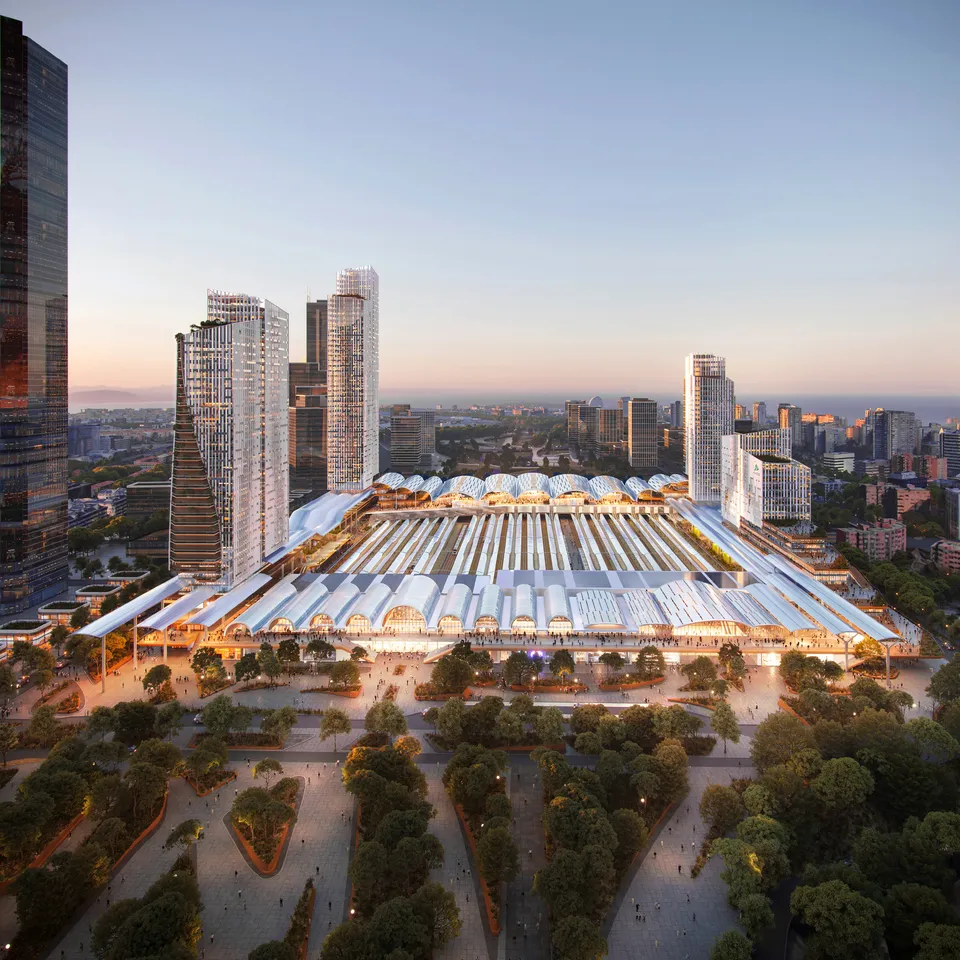 Aerial view of a modern train station with multiple platforms, surrounded by tall buildings and a park filled with trees at sunset.