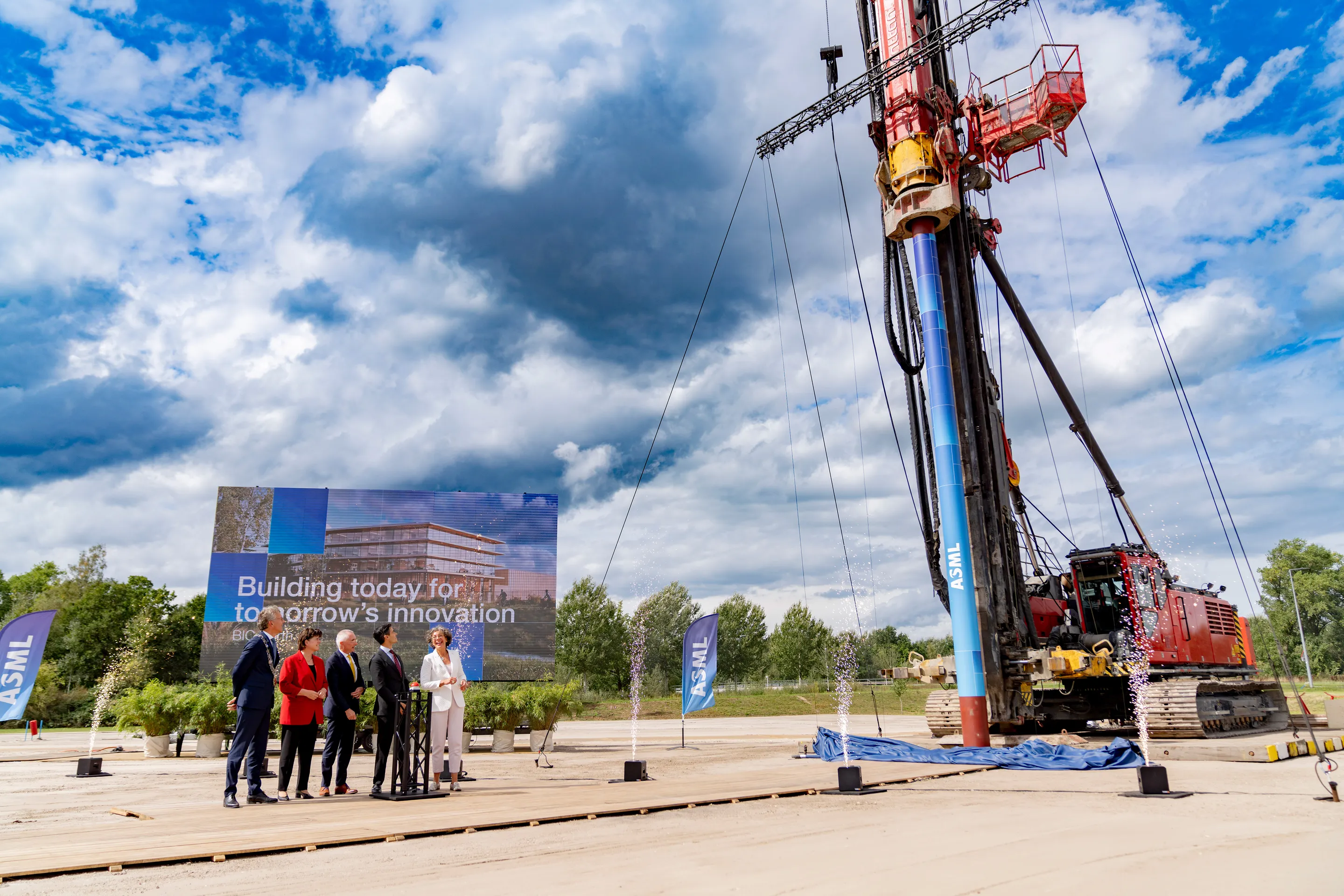 A group of people stands on a platform next to a large drilling machine and a sign that reads "Building today for tomorrow's innovation" with fireworks.
